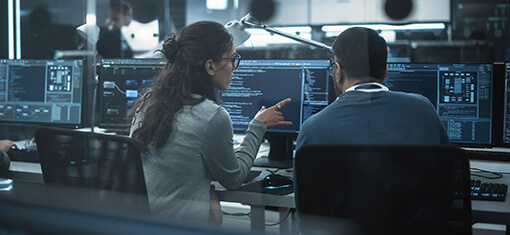 Eine Frau und ein Mann schauen auf zwei Computerbildschirme, die Programmcode anzeigen.