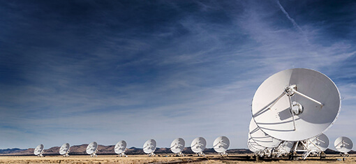 Ein Feld von Satellitenschüsseln in der Steppe mit weit entfernter Gebirgskette im Hintergrund und blauem Himmel, durchzogen von weißen Wolken.