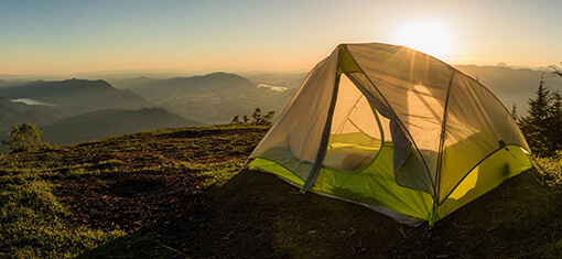 Ein Campingzelt auf einem Hügel mit Blick auf ein grünes Tal im Morgengrauen. Im Hintergrund geht die Sonne auf.