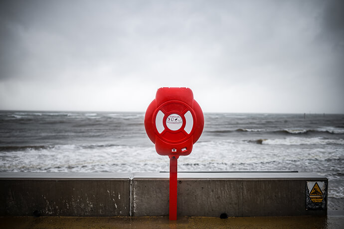 A gray, slightly stormy day at the beach. In the foreground, a red life ring hangs from a holder to be readily available in case of emergencies.