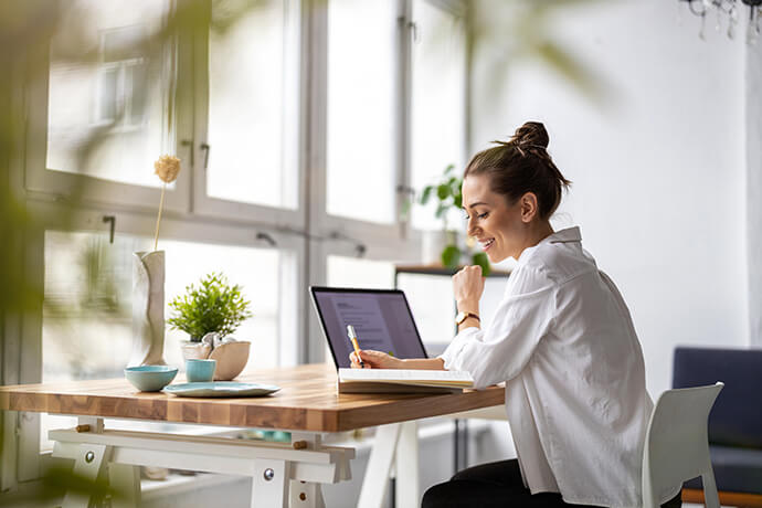 Eine Frau arbeitet entspannt zu Hause am Frühstückstisch. Sie arbeitet mit einem Computer und schreibt Notizen in ein Heft.